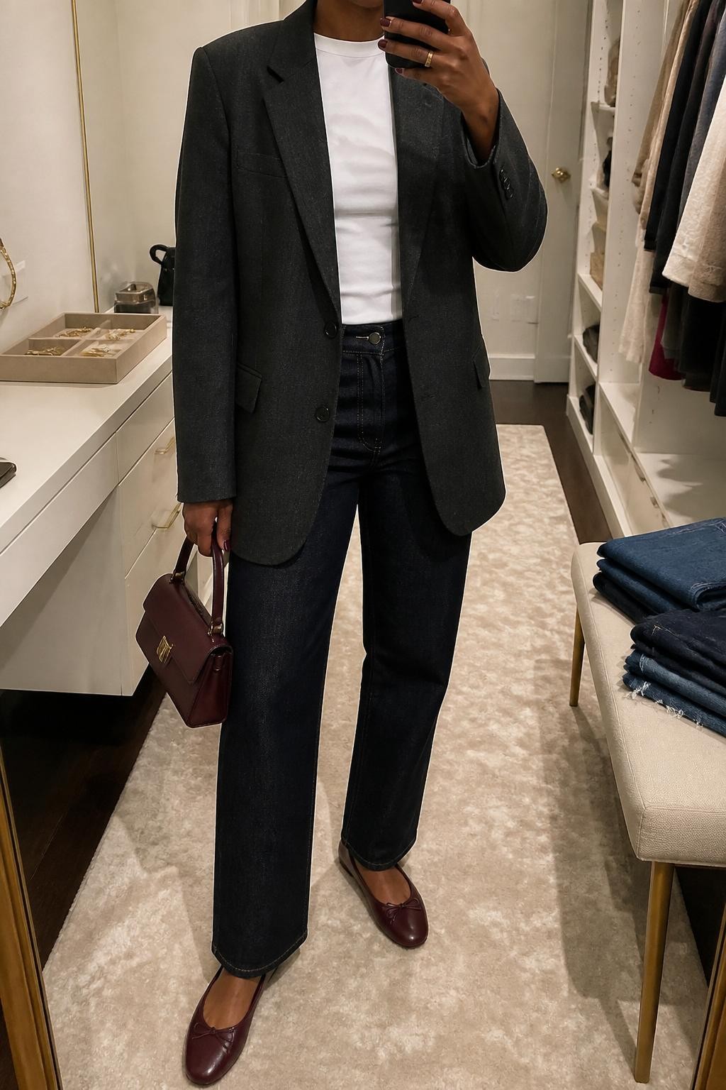 mirror selfie of a Black woman wearing an oversized charcoal wool blazer, fitted white crew-neck tee, dark indigo straight jeans, burgundy ballet flats, and a small burgundy leather handbag, clearly showing the same clothing items, colors, fabrics, and accessories listed in the Outfit Pieces section, holding a phone in front of a mirror, cropped at NECK so the face does not show, with a relaxed casual selfie pose, in a walk-in closet mirror, soft indoor lighting, including a jewelry tray on the vanity and folded denim jeans on a bench, latest iPhone photo quality.
