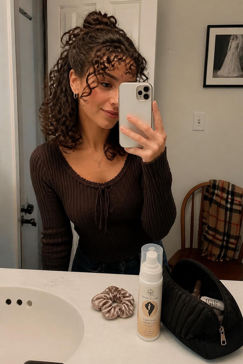 Photo of a woman wearing shoulder-length curls styled into a relaxed half-up top knot with loose curly fringe and softly defined lengths, candid mirror selfie in a bathroom vanity mirror, warm soft lighting, silk scrunchie and curl foam resting beside a makeup pouch, small hoop earrings peeking through and a plaid scarf draped over a chair, realistic everyday styling moment, latest iPhone photo quality.