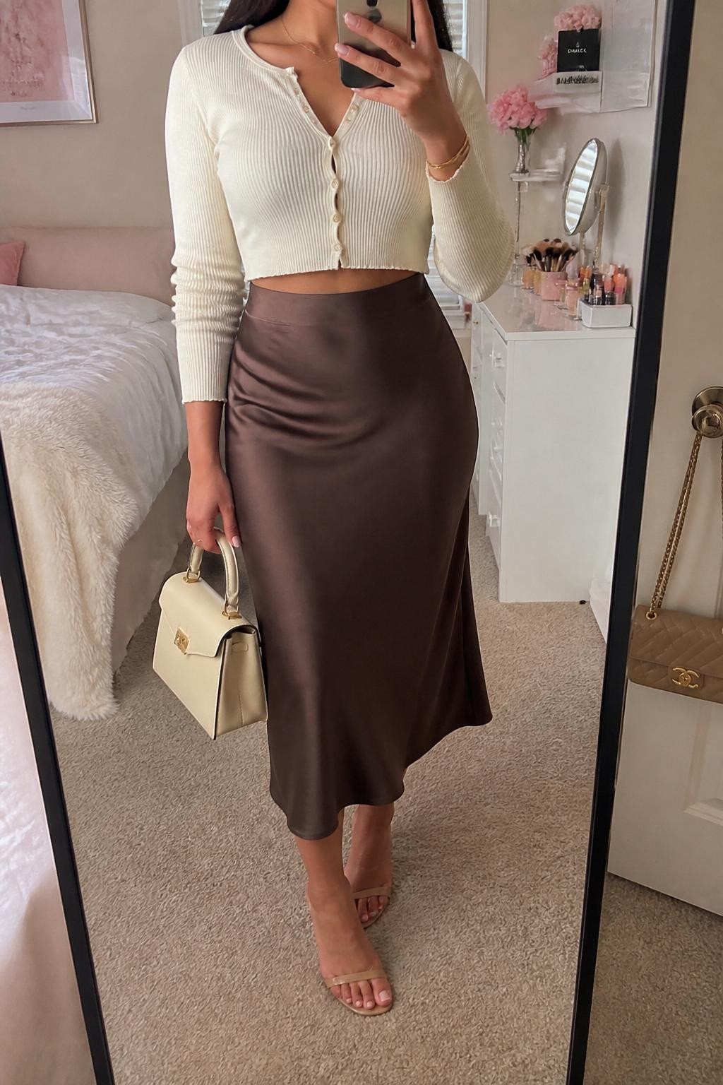 mirror selfie of a Latina woman wearing a cream cropped cardigan, brown satin midi skirt, nude heeled sandals, gold bracelet, and a cream top-handle bag, holding a phone in front of a mirror, cropped at NECK so the face does not show, relaxed casual selfie pose, in a feminine-themed bedroom with natural window light, including a makeup brush on the vanity and a handbag hanging on a door handle, realistic latest iPhone photo quality.