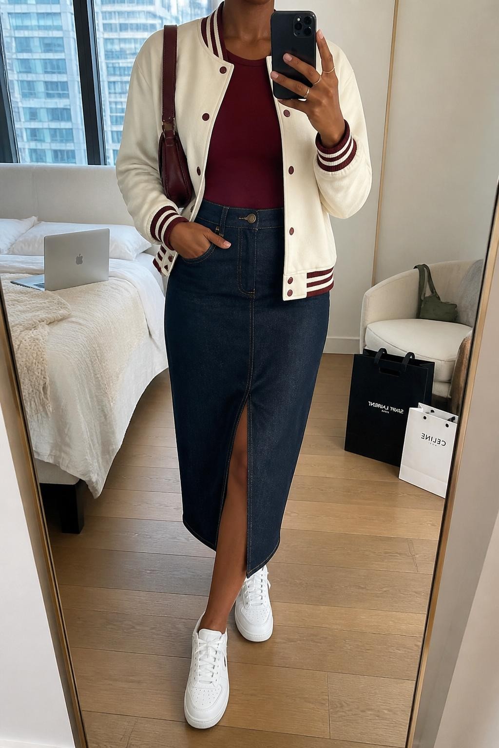 mirror selfie of a Black woman wearing a cream varsity jacket with burgundy trim, fitted burgundy crew-neck top, dark-wash denim midi skirt with a front slit, white leather sneakers, and a burgundy shoulder bag, clearly showing the same clothing items, colors, fabrics, footwear, and accessories listed in the Outfit Pieces section, holding a phone in front of a mirror, cropped at NECK so the face does not show, with a relaxed casual selfie pose, in a stylish city apartment, natural window light, including a laptop on the bed and shopping bags beside a chair, latest iPhone photo quality.