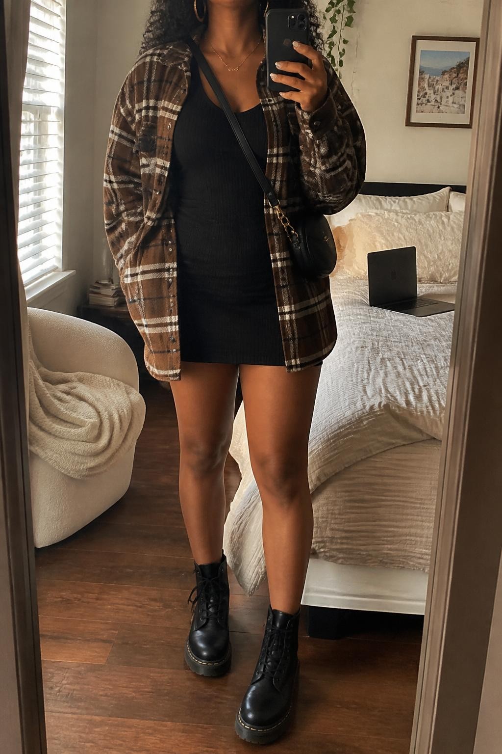 mirror selfie of an Afro-Latina woman wearing a brown plaid oversized shacket over a black ribbed mini dress, black combat boots, gold hoop earrings, and a small black crossbody bag, holding a phone in front of a mirror, cropped at NECK so the face does not show, relaxed casual selfie pose, in a cozy bedroom mirror setup with natural window light, including a folded blanket on a chair and a laptop on the bed, realistic latest iPhone photo quality.