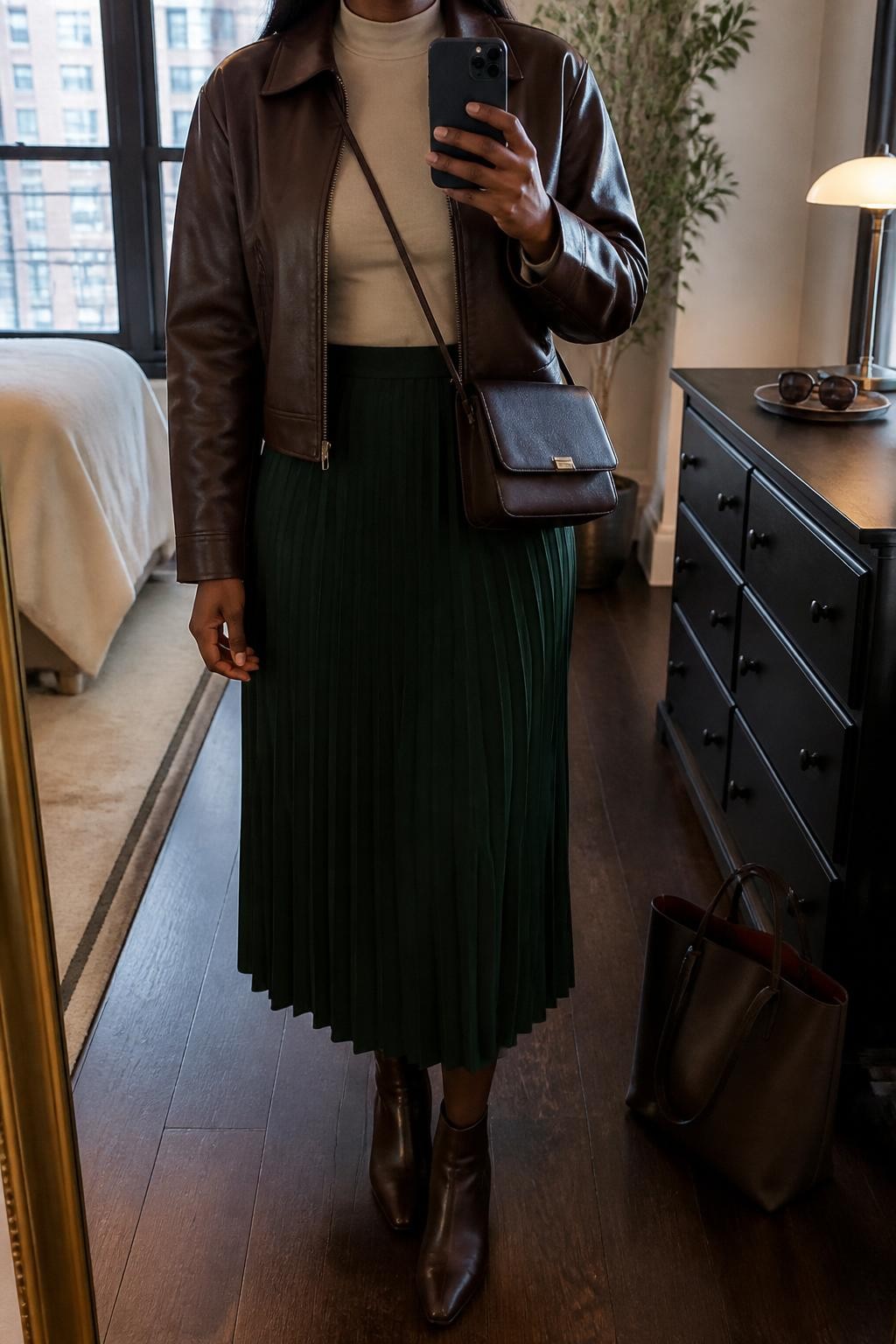 Mirror selfie of a Black woman with deep brown skin wearing a cropped espresso brown leather jacket over a fitted beige mock-neck top, a forest green pleated midi skirt, dark brown leather ankle boots, and a compact espresso leather crossbody bag, clearly showing the exact clothing items, colors, textures, and accessories listed, holding a latest-model iPhone in front of a mirror, cropped at the neck so the face does not show, relaxed casual selfie pose, in a stylish city apartment with warm moody lighting, a tote bag resting on the floor and sunglasses placed on a dresser, realistic casual setting, latest iPhone photo quality.