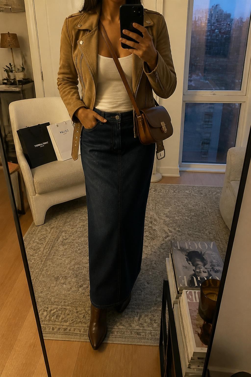 mirror selfie of a Latina woman wearing a tan suede moto jacket, white fitted tank top, dark denim maxi skirt, brown western ankle boots, and a cognac leather crossbody bag, holding a phone in front of a mirror, cropped at NECK so the face does not show, relaxed casual selfie pose, in a stylish city apartment with warm moody lighting, including shopping bags beside a chair and a stack of fashion magazines on a side table, realistic latest iPhone photo quality.