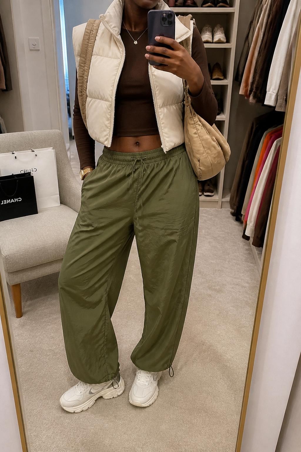 mirror selfie of a Black woman wearing a cropped cream puffer vest, fitted chocolate brown long-sleeve top, olive green parachute pants, chunky white sneakers, and a beige nylon shoulder bag, clearly showing the same clothing items, colors, fabrics, and accessories listed in the Outfit Pieces section, holding a phone in front of a mirror, cropped at NECK so the face does not show, with a relaxed casual selfie pose, in a walk-in closet mirror, soft indoor lighting, including shopping bags beside a chair and an open closet with clothes visible, latest iPhone photo quality.