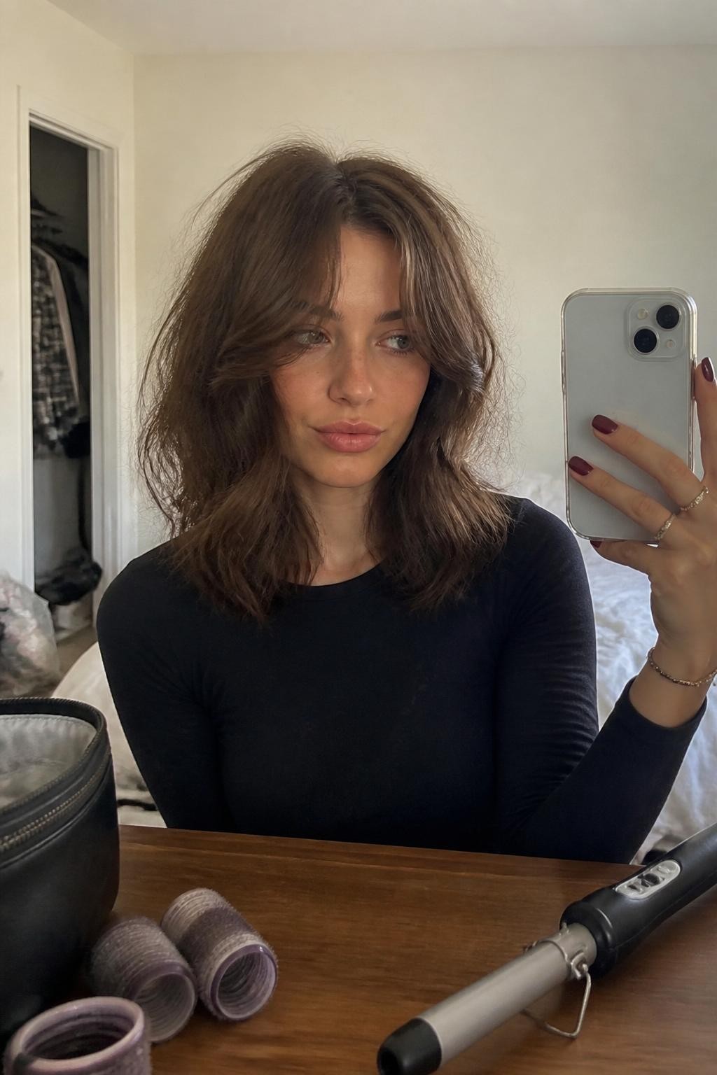 Photo of a woman wearing collarbone-length tousled waves with long curtain fringe sweeping away from the center of her face, candid mirror selfie in a softly lit bedroom, natural window light, curling wand on the dresser and two large rollers resting beside a makeup bag, a plaid shirt visible inside a slightly open closet, realistic second-day texture and loose flyaways, latest iPhone photo quality.