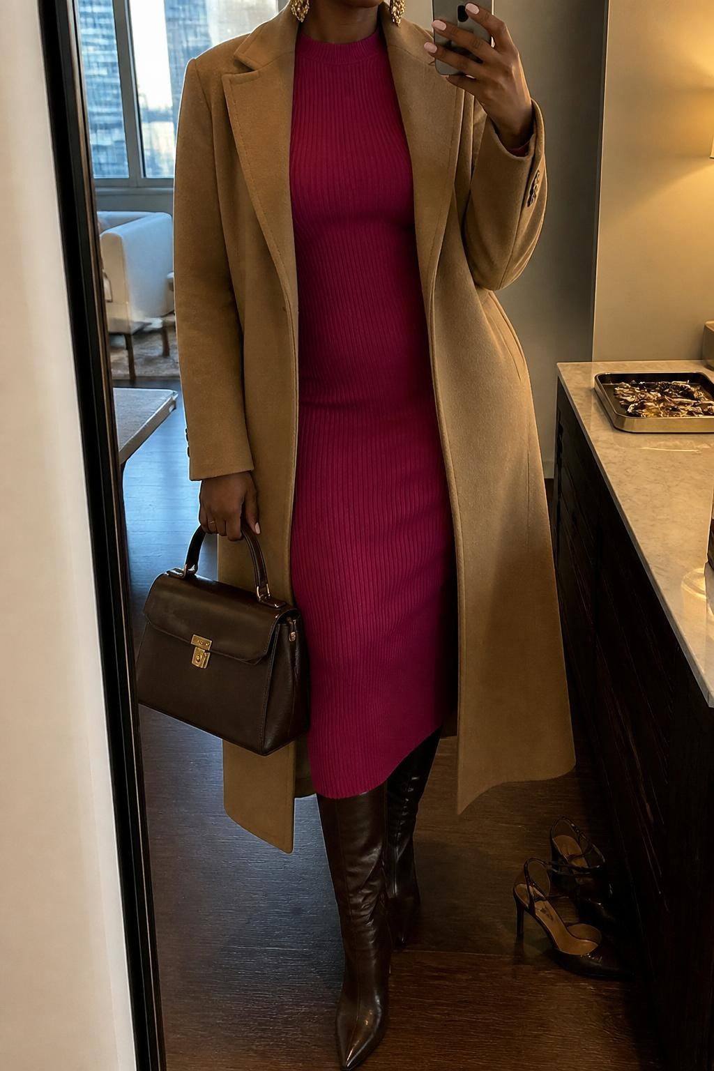 mirror selfie of a Black woman wearing a fitted fuchsia ribbed midi dress under a long camel wool coat, chocolate brown leather knee-high boots, gold statement earrings, and a structured dark brown handbag, clearly showing the same clothing items, colors, fabrics, and accessories listed in the Outfit Pieces section, holding a phone in front of a mirror, cropped at NECK so the face does not show, with a relaxed casual selfie pose, in a stylish city apartment, warm moody lighting, including a jewelry tray on the vanity and a pair of heels on the floor nearby, latest iPhone photo quality.