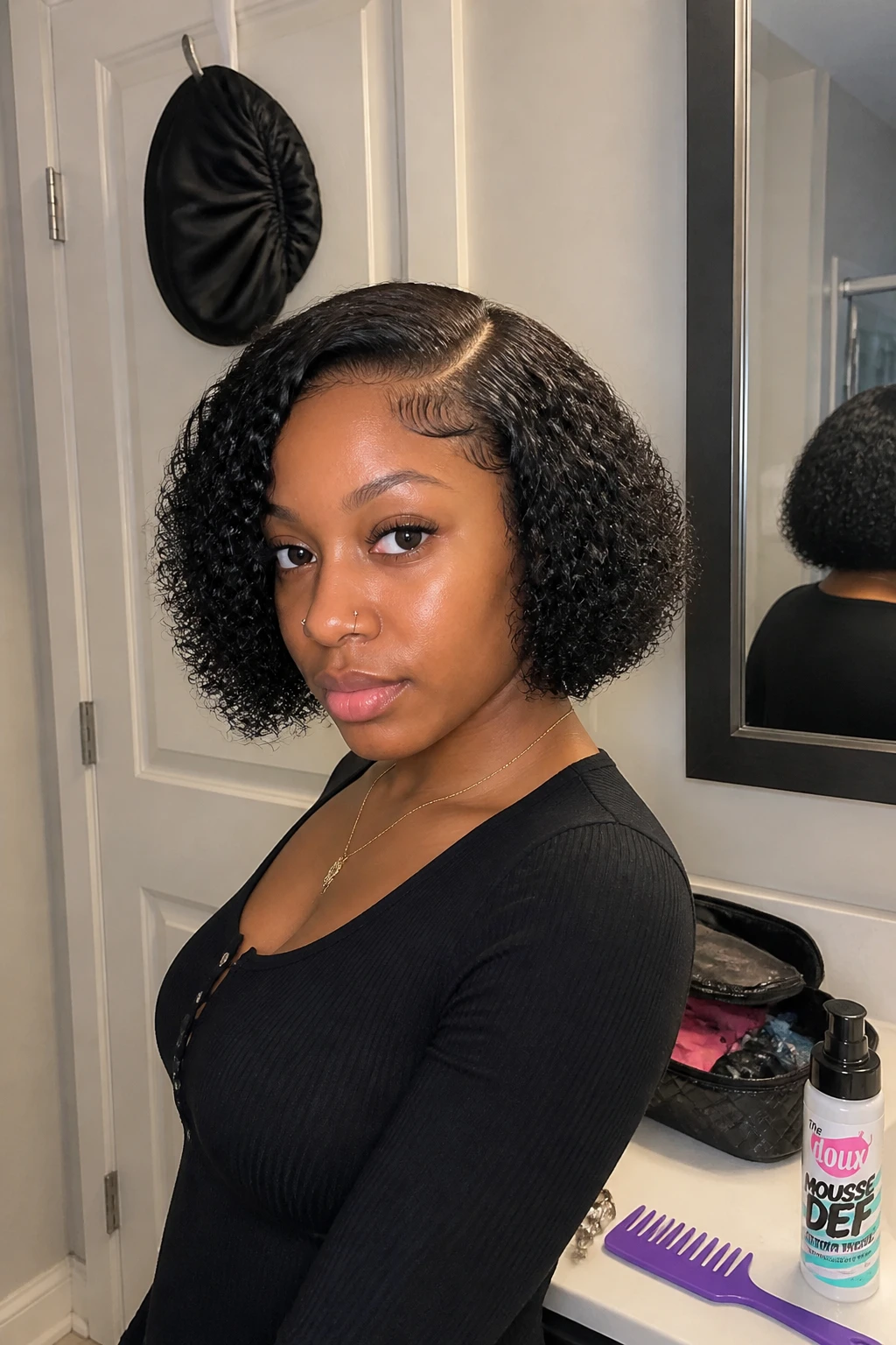 Image prompt
Photo of a Black woman wearing a chin-length curly sew-in bob with a softly rounded shape, springy ringlets, a clean invisible part, and lightly shaped ends around the jaw, front three-quarter view beside a bathroom vanity mirror, soft indoor lighting, containing a small lived-in detail such as a wide-tooth comb and curl mousse on the counter, a makeup bag nearby, and a satin bonnet hanging from the door, realistic getting-ready moment, latest iPhone photo quality.