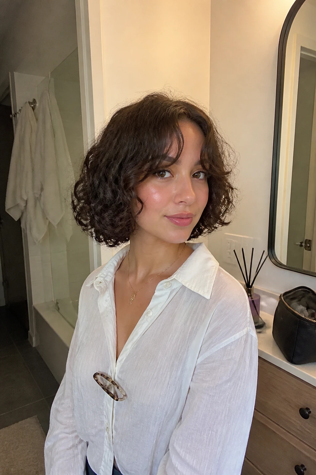 Image prompt

Photo of a woman wearing a chin-length curly bob with soft curtain bangs opening around the forehead and springy curls framing the jaw, front three-quarter view beside a bathroom vanity mirror, soft indoor lighting, containing a small lived-in detail such as a diffuser on the counter, a makeup bag nearby, and a hair clip clipped to her blouse collar, realistic getting-ready moment, latest iPhone photo quality.