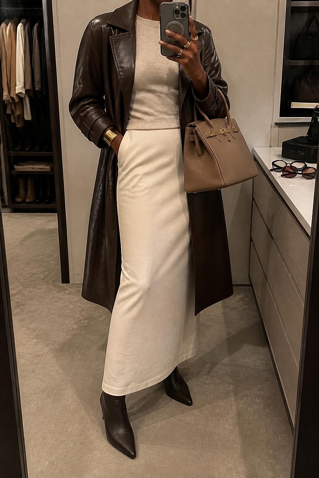 mirror selfie of a Black woman wearing the exact outfit described above, clearly showing an espresso brown leather trench jacket layered over a fitted oatmeal knit top, cream column maxi skirt, brown kitten-heel ankle boots, a tan structured handbag, and gold cuff bracelet, holding a phone in front of a mirror, cropped at NECK so the face does not show, with a relaxed casual selfie pose, in a walk-in closet mirror, warm moody lighting, with an open closet with clothes visible and sunglasses placed on a dresser, latest iPhone photo quality.