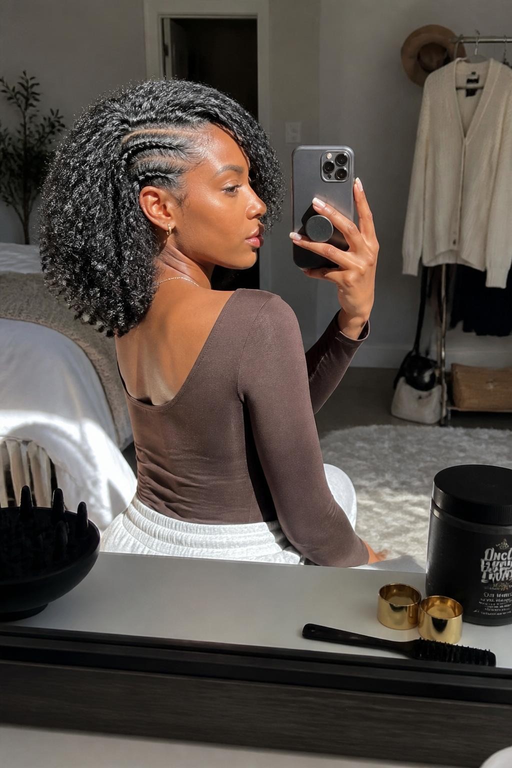 Photo of a Black woman wearing a shoulder-length defined wash-and-go with a deep side part, springy curls, and two slim flat braids following the smaller side of the head, side-profile mirror selfie in a sun-drenched bedroom, natural window light, curl gel and diffuser resting on the dresser, gold cuffs beside an edge brush and a cream cardigan visible behind her, realistic shrinkage and soft frizz, latest iPhone photo quality.