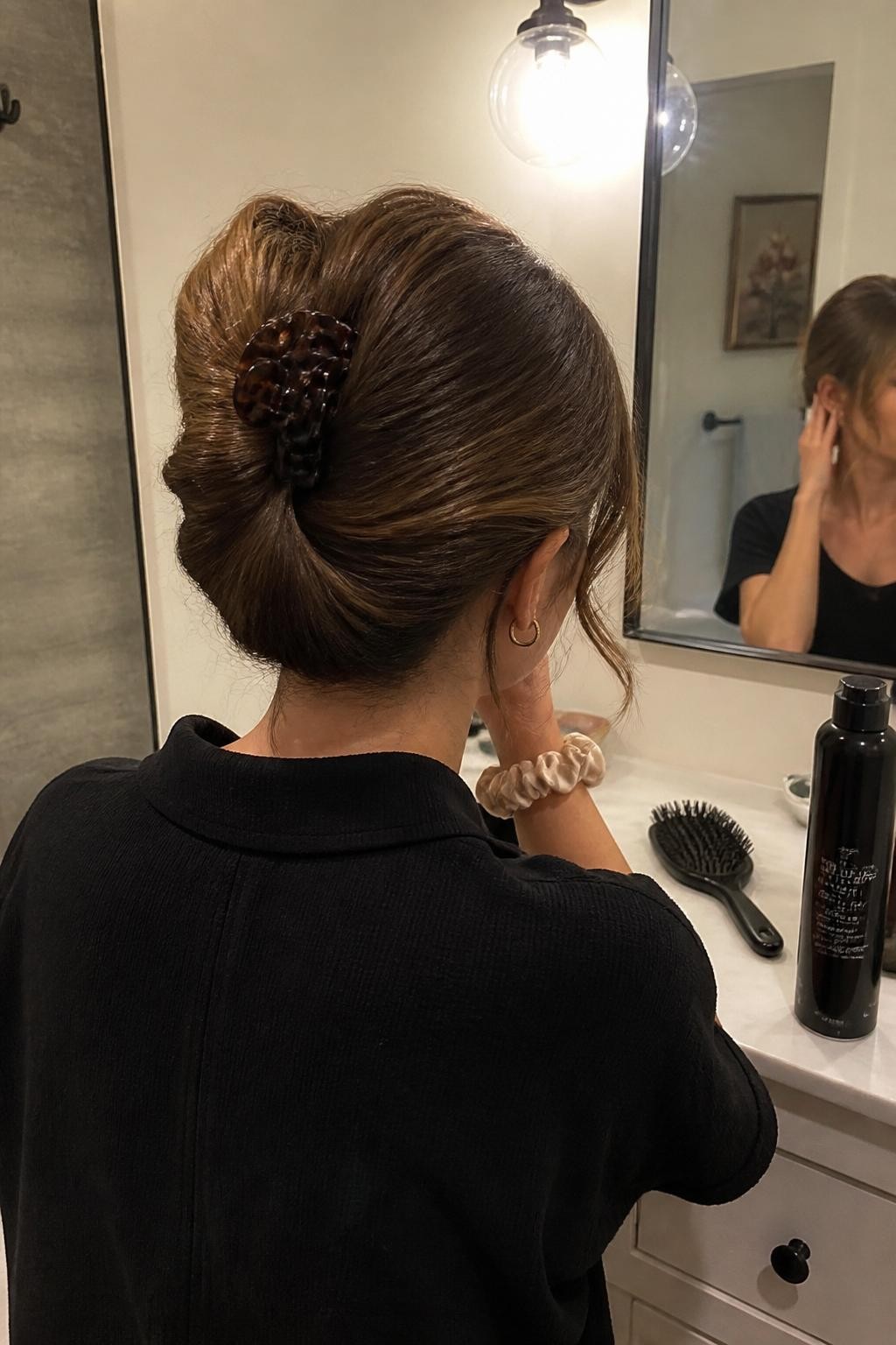 Photo of a woman wearing a medium-length French roll with softly folded ends held by a large tortoiseshell claw clip, back-of-head view at a bathroom vanity mirror, soft indoor lighting, hairbrush and dry-texture spray on the counter, loose tendrils near the ears and a silk scrunchie around her wrist, realistic slightly imperfect twist, latest iPhone photo quality.