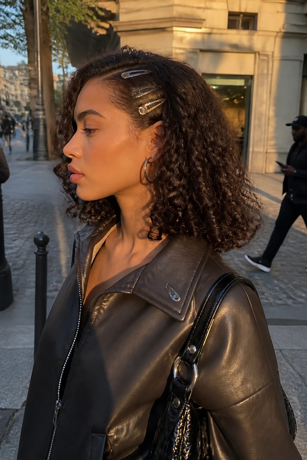 Image prompt
Photo of a woman wearing shoulder-length curly hair with a deep side part, softly flipped ends, and three small metallic clips placed above one ear, side profile view against a softly blurred city street background, golden hour glow, containing a small lived-in detail such as hoop earrings, a glossy mini handbag, and one spare clip attached to her jacket collar, realistic candid Y2K-inspired photograph, latest iPhone photo quality.