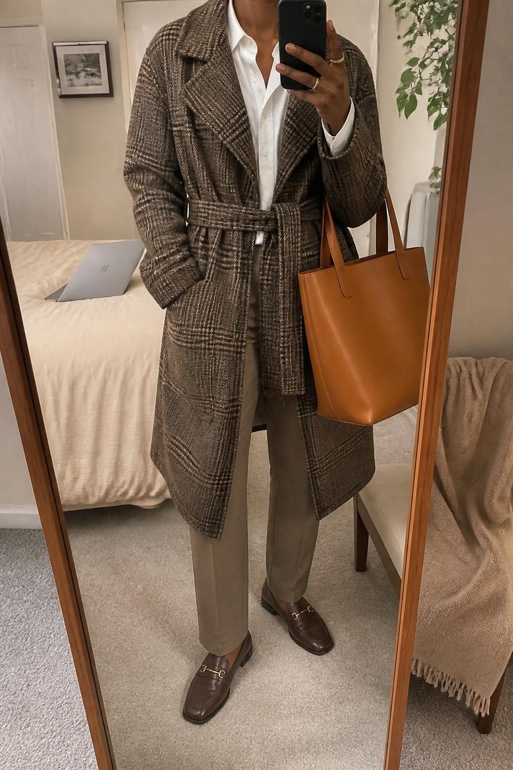 mirror selfie of a Black woman wearing a brown plaid wrap coat, white cotton button-down shirt, taupe straight-leg pants, dark brown loafers, and tan leather tote, clearly showing the same clothing items, colors, fabrics, and accessories listed in the Outfit Pieces section, holding a phone in front of a mirror, cropped at NECK so the face does not show, with a relaxed casual selfie pose, in a cozy bedroom mirror setup, soft indoor lighting, including a laptop or tablet on the bed and a folded blanket on a chair, latest iPhone photo quality.