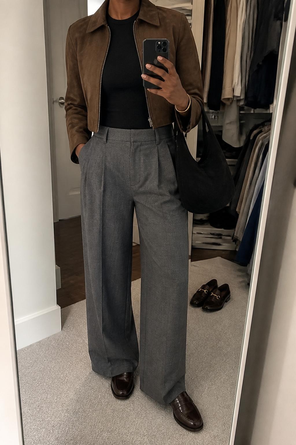 Mirror selfie of a Black woman with deep brown skin wearing a cropped chocolate brown suede jacket over a fitted black crew-neck knit top, high-waisted gray wide-leg pleated trousers, dark brown leather loafers, and a soft black suede hobo bag, clearly showing the exact clothing items, colors, fabrics, and accessories listed, holding a latest-model iPhone in front of a mirror, cropped at the neck so the face does not show, relaxed casual selfie pose, in a walk-in closet mirror with soft indoor lighting, an open closet with clothes visible and a pair of loafers on the floor nearby, realistic uncluttered setting, latest iPhone photo quality.