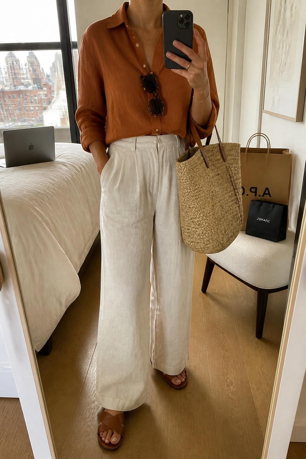 mirror selfie of a woman wearing a rust orange button-up shirt, cream linen-blend wide-leg pants, brown slide sandals, a straw tote bag, and tortoiseshell sunglasses, clearly showing the same clothing items, colors, fabrics, and accessories listed in the Outfit Pieces section, holding a phone in front of a mirror, cropped at NECK so the face does not show, with a relaxed casual selfie pose, in a stylish city apartment, natural window light, including a laptop on the bed and shopping bags beside a chair, latest iPhone photo quality.
