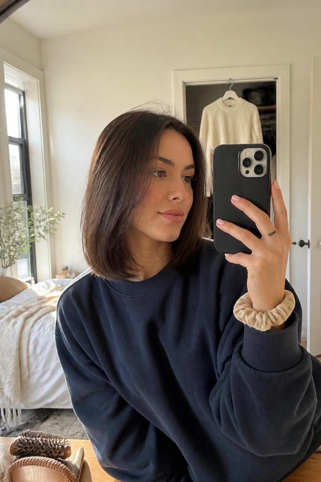 Photo of a woman wearing a collarbone-length soft blunt lob with a center part and smooth ends gently curved inward, candid mirror selfie taken slightly from the side, sun-drenched bedroom with a cream sweater hanging from a slightly open closet, natural window light, round brush on the dresser and a silk scrunchie around her wrist, realistic flyaways and natural hair movement, latest iPhone photo quality.