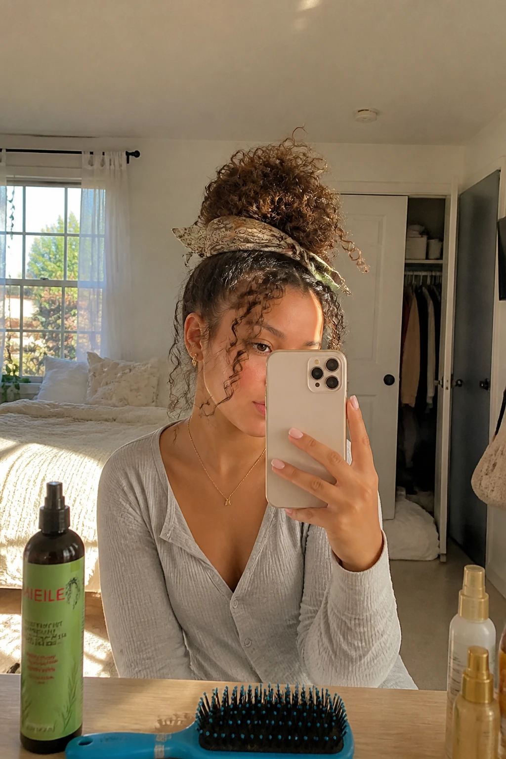 Image prompt

Photo of a woman wearing a high curly bun wrapped at the base with a narrow printed satin scarf, soft curl ends visible around the bun, and a few face-framing pieces, candid mirror selfie in a sun-drenched bedroom, natural window light, containing a small lived-in detail such as a curl refresher bottle on the vanity, a hairbrush nearby, and a slightly open closet behind her, realistic bad hair day getting-ready moment, latest iPhone photo quality.