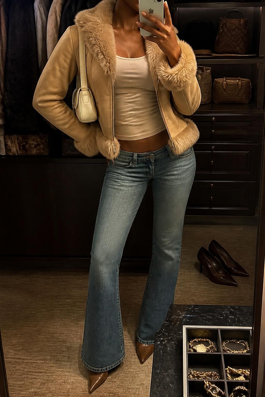 mirror selfie of a Black woman wearing a tan faux fur trim jacket, a soft beige fitted tank top, low-rise medium-wash flare jeans, pointed brown heeled boots, and a small cream shoulder bag, clearly showing the same clothing items, colors, fabrics, and accessories listed in the Outfit Pieces section, holding a phone in front of a mirror, cropped at NECK so the face does not show, with a relaxed casual selfie pose, in a walk-in closet mirror, warm moody lighting, including a jewelry tray on the vanity and a pair of heels on the floor nearby, latest iPhone photo quality.