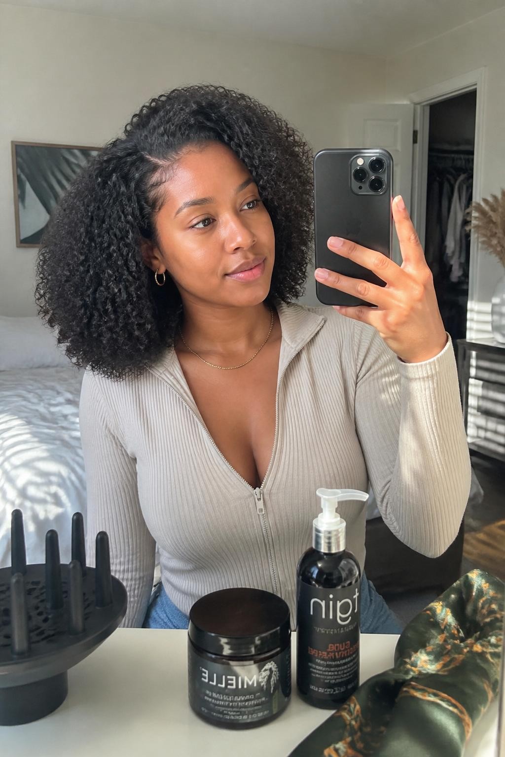 Image prompt: Photo of a Black woman wearing a defined shoulder-length wash-and-go with springy curls, a deep side part, and lifted roots, candid mirror selfie, sun-drenched bedroom setting, natural window light, diffuser and curl cream visible on the dresser, satin scarf loosely placed nearby and a slightly open closet behind her, realistic natural-hair routine, latest iPhone photo quality.