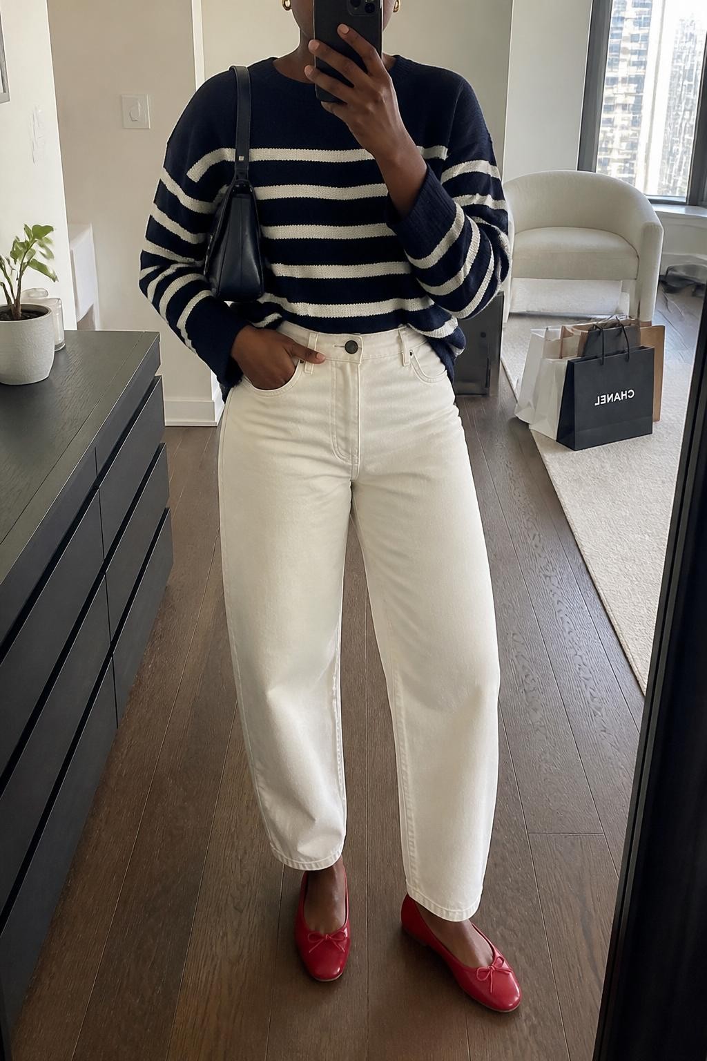mirror selfie of a Black woman wearing the exact outfit described above, clearly showing a navy and cream striped sweater, cream barrel jeans, red ballet flats, a navy leather shoulder bag, and small gold earrings, holding a phone in front of a mirror, cropped at NECK so the face does not show, with a relaxed casual selfie pose, in a stylish city apartment, natural window light, with shopping bags beside a chair and a small plant on a dresser, latest iPhone photo quality.