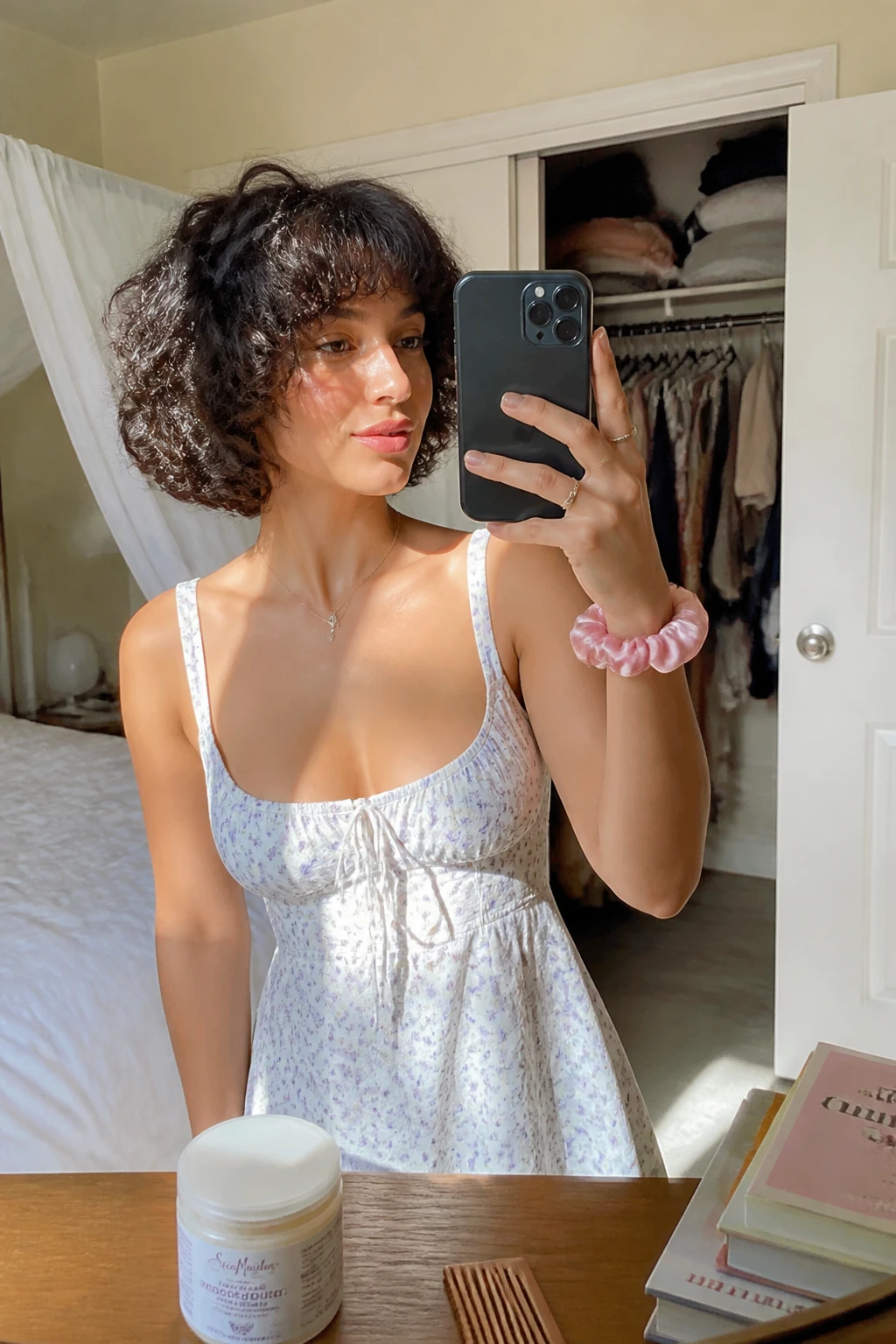 Image prompt

Photo of a woman wearing a short layered curly bob with soft rounded volume, defined curls, and light face-framing pieces around the cheeks, candid mirror selfie in a sun-drenched bedroom, natural window light, containing a small lived-in detail such as a curl cream jar on the dresser, a silk scrunchie on her wrist, and a slightly open closet behind her, realistic getting-ready moment, latest iPhone photo quality.