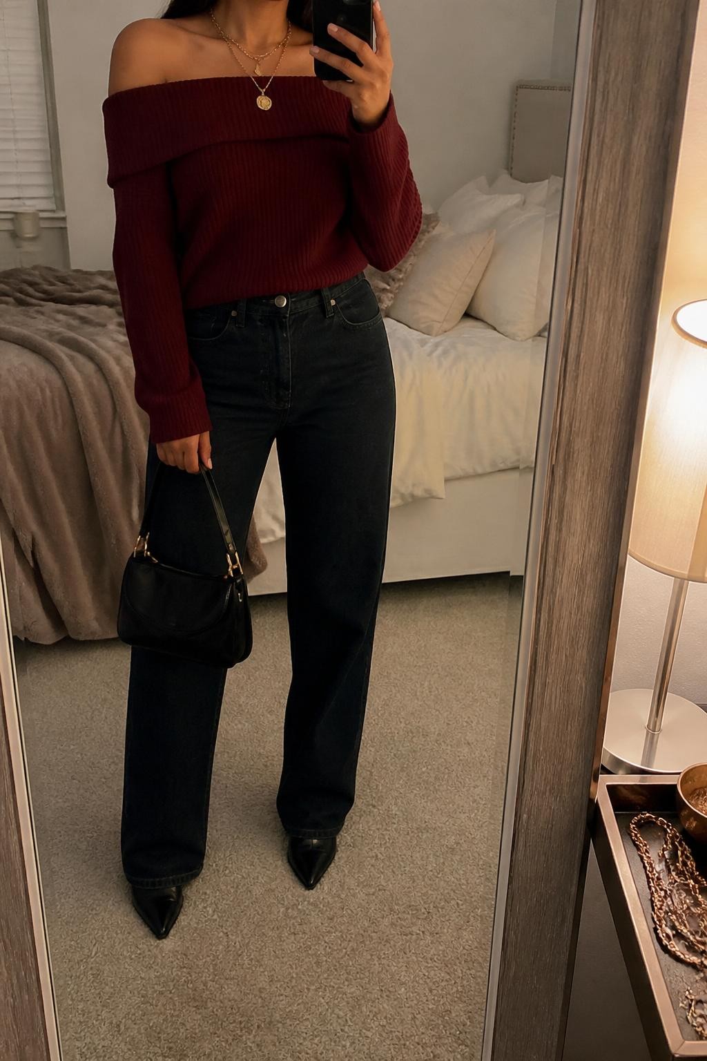 mirror selfie of a Latina woman wearing a burgundy off-shoulder knit sweater, dark wash high-waisted straight jeans, black pointed ankle boots, gold layered necklace, and a black mini handbag, holding a phone in front of a mirror, cropped at NECK so the face does not show, relaxed casual selfie pose, in a cozy bedroom mirror setup with warm moody lighting, including soft pillows on an unmade bed and a jewelry tray on the vanity, realistic latest iPhone photo quality.