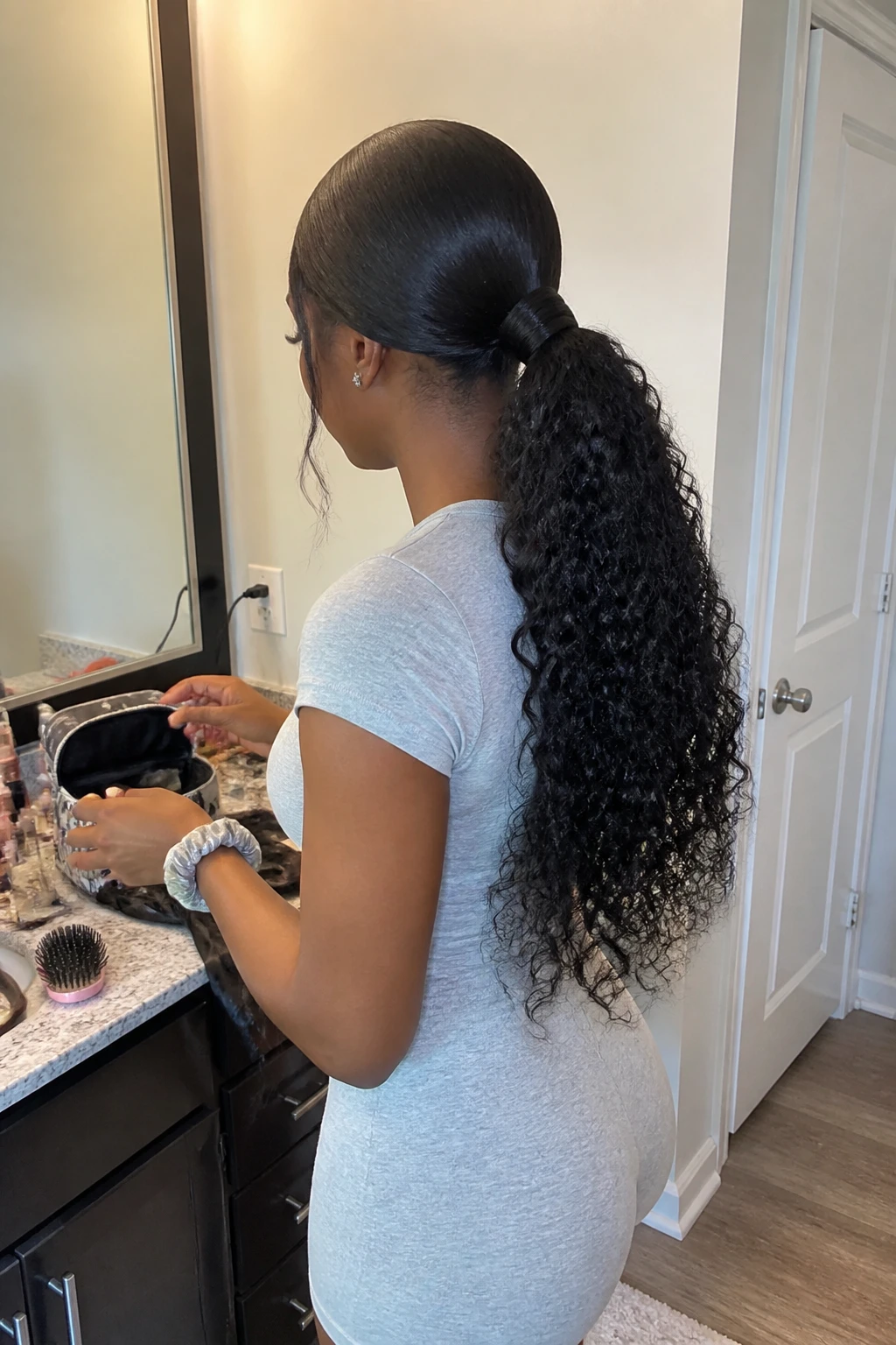 Image prompt
Photo of a Black woman wearing long curly sew-in hair in a low ponytail with a clean middle part, softly smoothed crown, wrapped hair section around the elastic, and full defined curls falling down her back, back three-quarter view beside a bathroom vanity, soft indoor lighting, containing a small lived-in detail such as a hairbrush on the counter, a satin scarf near an open makeup bag, and a silk scrunchie on her wrist, realistic getting-ready moment, latest iPhone photo quality.