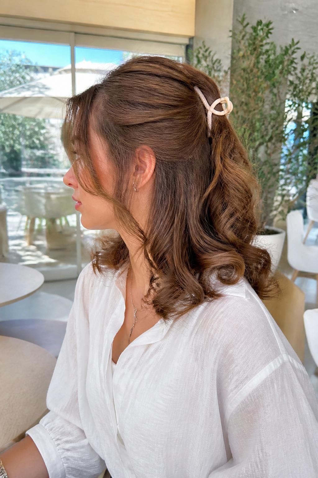 A realistic beauty photo of a woman with shoulder-length caramel brown curls styled in a half-up half-down mini bun secured with a small cream claw clip, lower curls left full and bouncy, soft curtain-like front tendrils, white blouse, airy café background, bright natural daylight, casual pretty hairstyle inspiration, high-resolution DSLR photo, no text, (no watermarks on images)