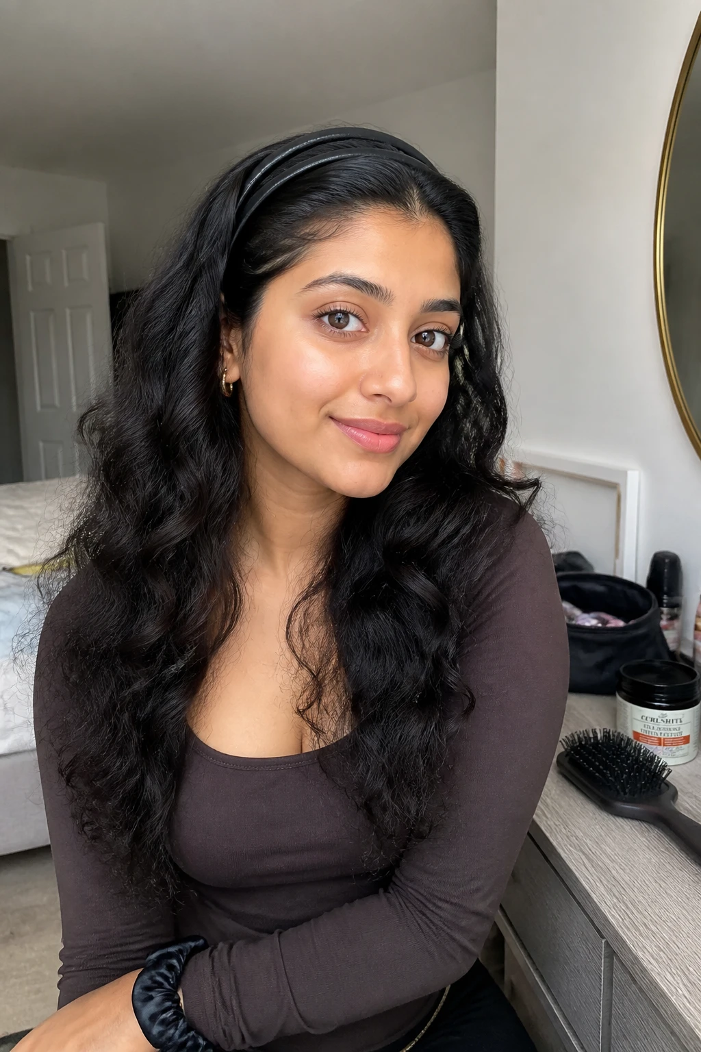 Image prompt

Photo of a South Asian woman wearing long loose curls with two slim black headbands placed closely together behind the hairline, front three-quarter view in a softly lit bedroom, natural window light, containing a small lived-in detail such as a curl cream jar near an open makeup bag, a silk scrunchie on her wrist, and a hairbrush resting on the dresser, realistic getting-ready moment, latest iPhone photo quality.