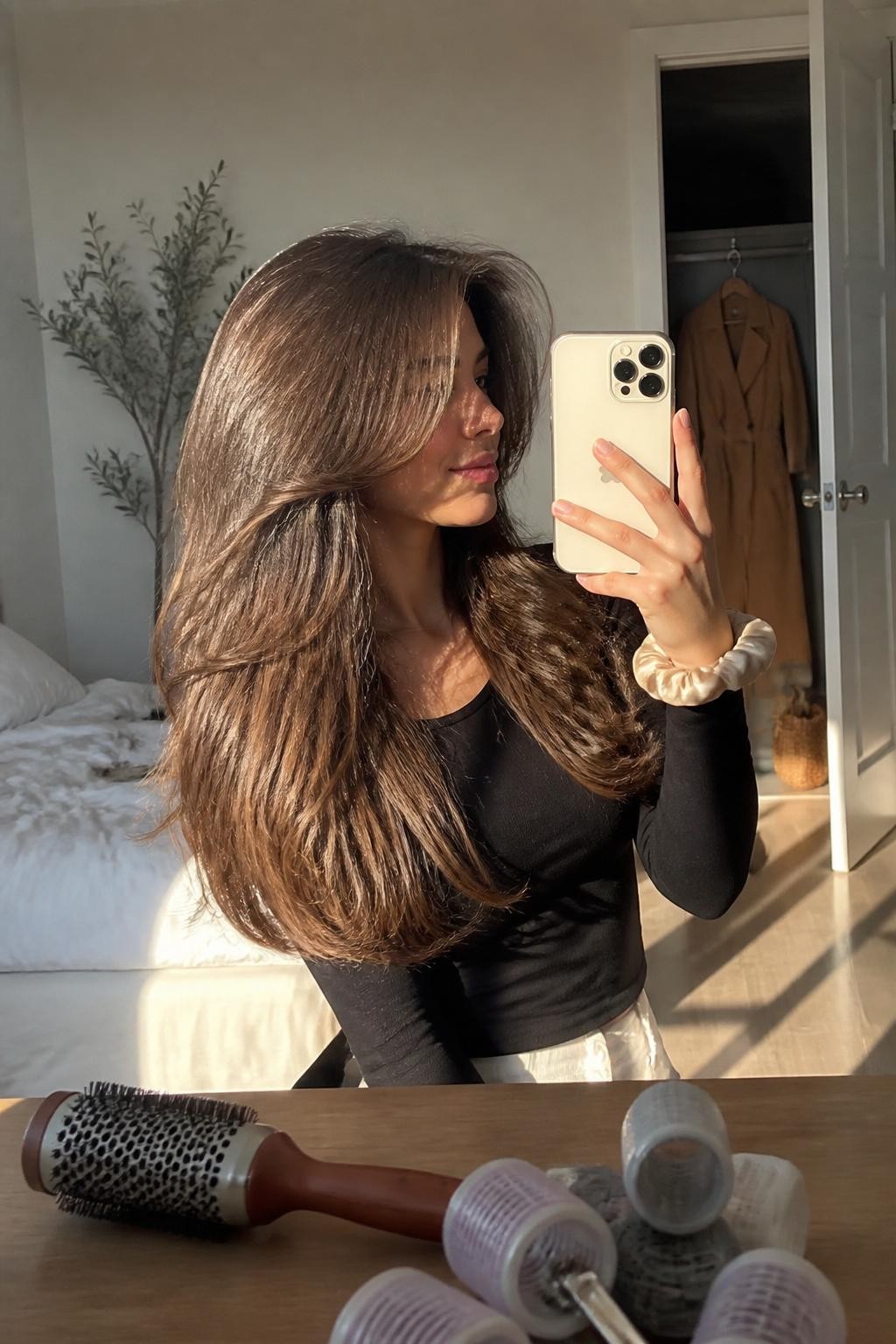 Photo of a woman wearing waist-length layered hair styled into a soft voluminous blowout with long curtain pieces and feathered ends sweeping away from the face, candid mirror selfie from a slight side angle in a sun-drenched bedroom, warm natural window light, round brush and several large rollers on the dresser, silk scrunchie around her wrist and a camel coat visible inside a slightly open closet, realistic movement and natural flyaways, latest iPhone photo quality.