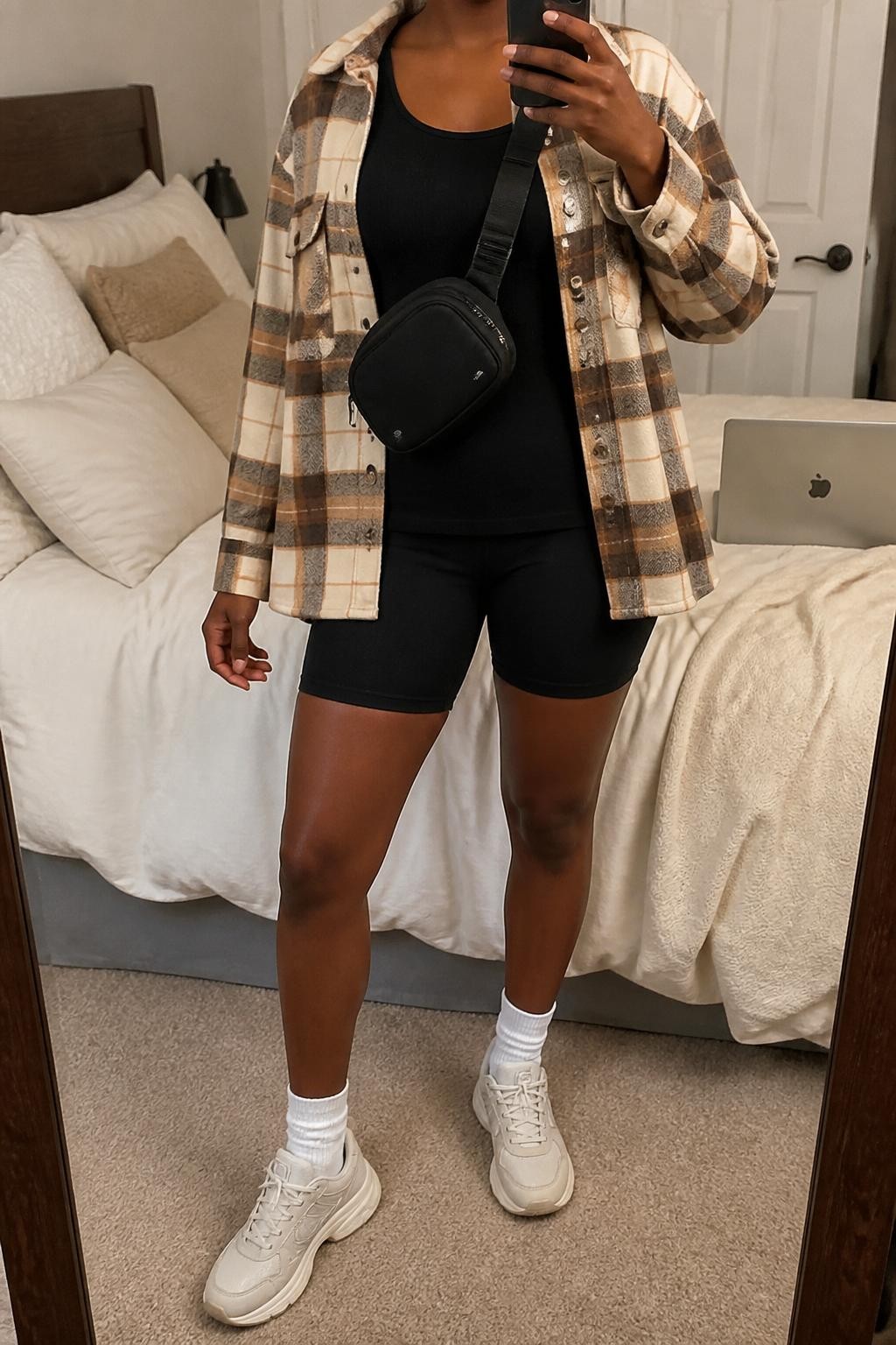 mirror selfie of a Black woman wearing a beige and brown plaid shacket, fitted black longline tank top, black bike shorts, white crew socks, chunky cream sneakers, and a black belt bag worn crossbody, clearly showing the same clothing items, colors, fabrics, and accessories listed in the Outfit Pieces section, holding a phone in front of a mirror, cropped at NECK so the face does not show, with a relaxed casual selfie pose, in a cozy bedroom mirror setup, soft indoor lighting, including soft pillows on an unmade bed and a laptop on the bed, latest iPhone photo quality.