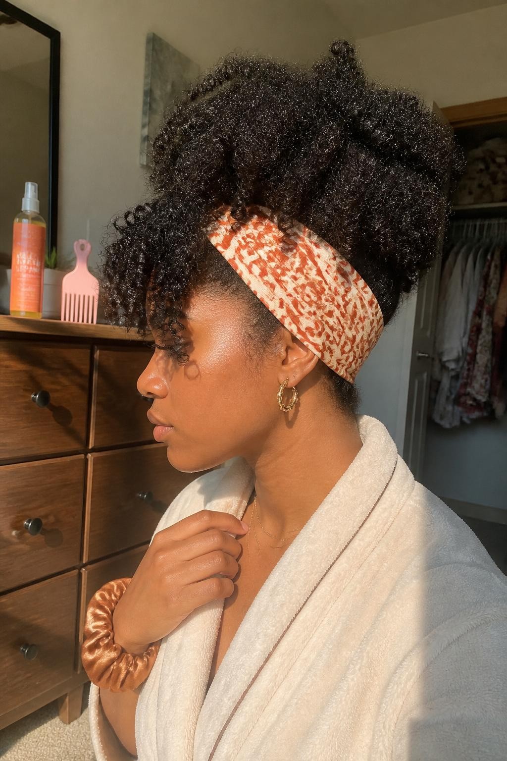 Image prompt: Photo of a Black woman wearing a high curly pineapple with defined coils spilling forward and a wide rust-and-cream printed headband, candid side-profile view, sun-drenched bedroom, warm natural window light, curl refresher bottle and hair pick on the dresser, silk scrunchie around her wrist, cozy robe and slightly open closet in the background, latest iPhone photo quality.