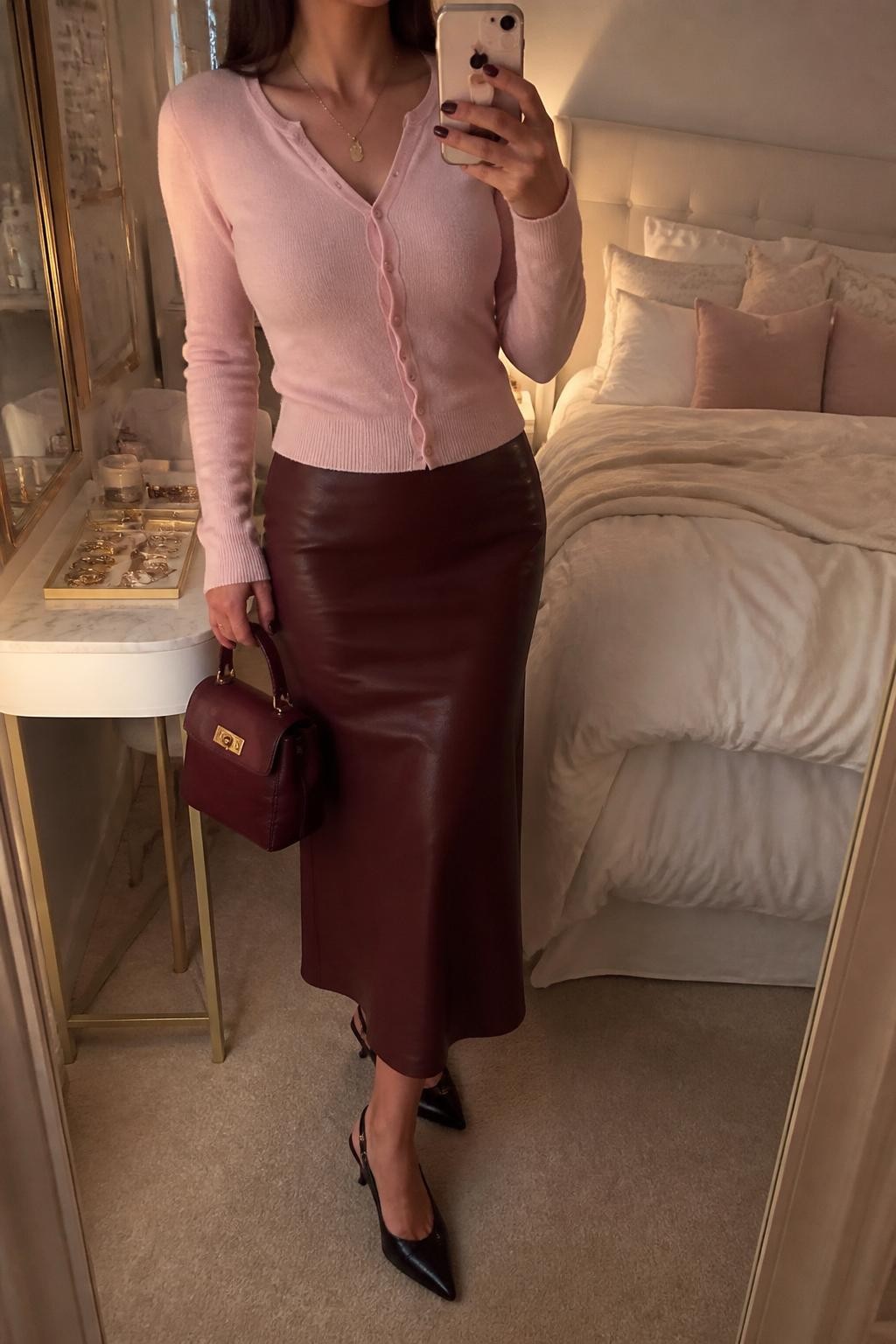 mirror selfie of a woman wearing a ballet pink fitted button-front cardigan, burgundy faux leather midi skirt, black pointed slingback heels, delicate gold pendant necklace, and a small burgundy top-handle bag, clearly showing the same clothing items, colors, fabrics, and accessories listed in the Outfit Pieces section, holding a phone in front of a mirror, cropped at NECK so the face does not show, with a relaxed casual selfie pose, in a feminine-themed bedroom, evening golden hour, including a jewelry tray on the vanity and soft pillows on an unmade bed, latest iPhone photo quality.